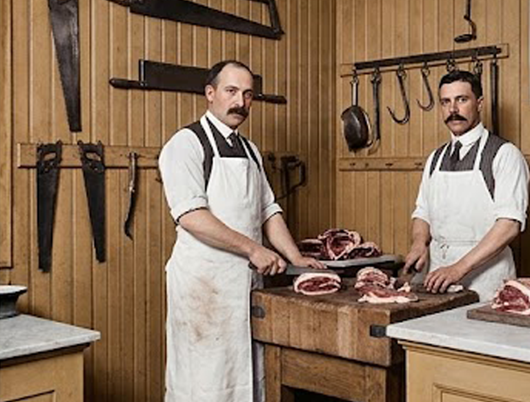 Neg - Restauro Affettatrici Berkel D'epoca E Volano | Wbs Maschi Two Butchers in White Aprons Cutting Meat on a Wooden Block Inside a Rustic Butcher Shop.