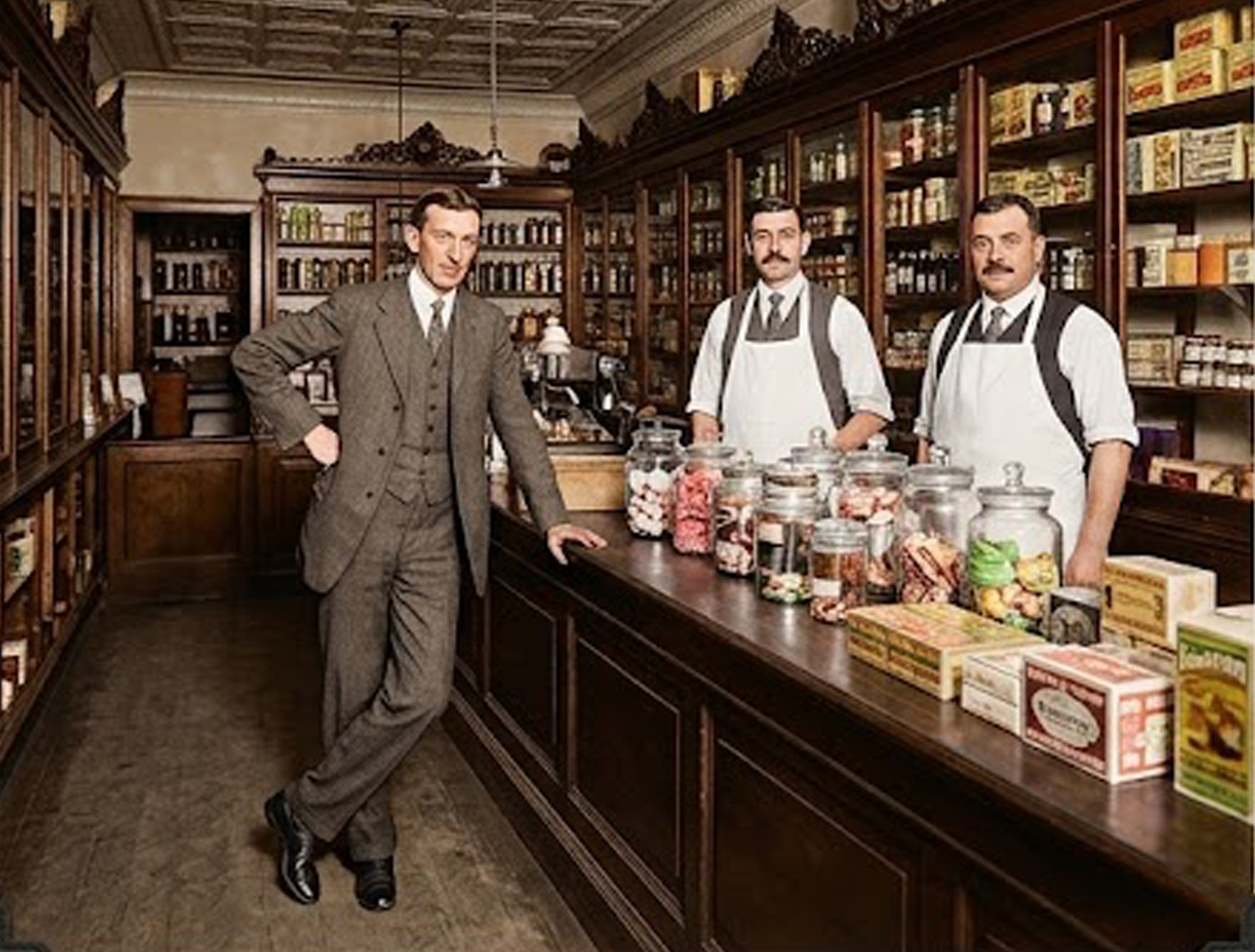 Negozio - Restauro Affettatrici Berkel D'epoca E Volano | Wbs Maschi Man in a Gray Suit Leans on a Dark Wooden Counter in a Vintage Apothecary; Two Clerks in White Aprons Stand Behind with Jars on Shelves.